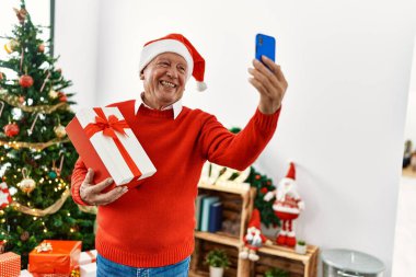 Yaşlı gri saçlı adam elinde noel hediyesiyle evde akıllı telefonuyla selfie çekiyor..