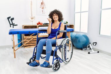 Fizyoterapi kliniğinde tekerlekli sandalyede oturan, gülümseyen, gülümseyen ve baş parmağını kaldırmış bir orta doğulu genç kadın.. 