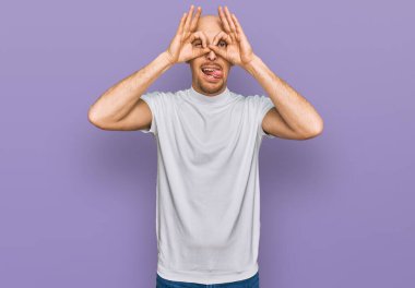 Sakallı, beyaz tişörtlü, kel bir adam dürbün gibi jest yapıyor. Dilini çıkarıyor, gözleri parmaklarından bakıyor. çılgın ifade. 