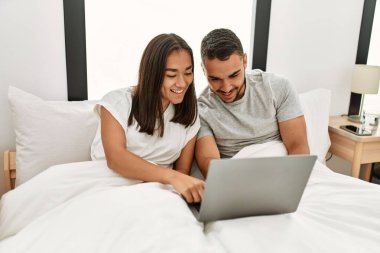 Yatak odasında dizüstü bilgisayar kullanarak mutlu gülümseyen genç Latin çift.
