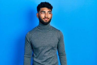 Balıkçı yaka kazak giyen sakallı Arap adam gülümseyip yan tarafa bakıyor ve düşünüyor.. 