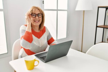 Orta yaşlı sarışın kadın dizüstü bilgisayar kullanıyor ve evde kahve içiyor..