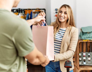 Genç İspanyol esnaf gülümseyerek giyim mağazasındaki müşteriye alışveriş çantası veriyor..