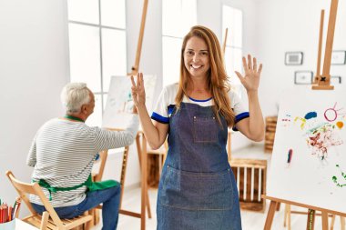 Hispanic woman wearing apron at art studio showing and pointing up with fingers number eight while smiling confident and happy. 