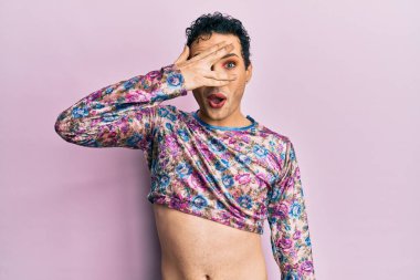 Handsome man wearing make up wearing fashion clothes peeking in shock covering face and eyes with hand, looking through fingers with embarrassed expression. 