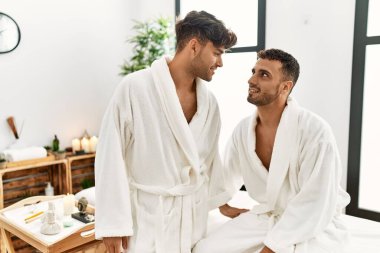 Two hispanic men couple wearing bathrobe sitting on massage table at beauty center