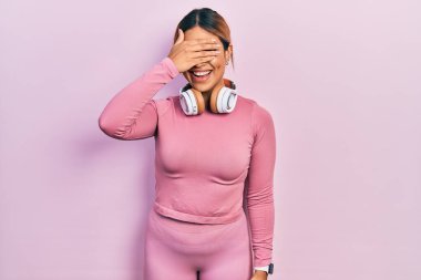 Güzel İspanyol bir kadın spor kıyafetleri giyiyor ve kulaklık takıyor. Gülümsüyor ve elleri yüzünde gülüyor. Şaşırtmak için gözleri örtüyor. kör kavram. 