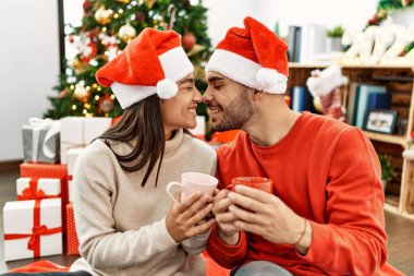 Mutlu gülümseyen ve kahve içen genç İspanyol çift evdeki Noel ağacının yanında yerde oturuyor..