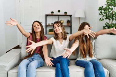 Üç İspanyol kızdan oluşan bir grup evdeki kanepede oturmuş kameraya bakıp sarılmak için kollarını açmış gülümsüyor. Mutluluğu kucaklayan neşeli bir ifade.. 