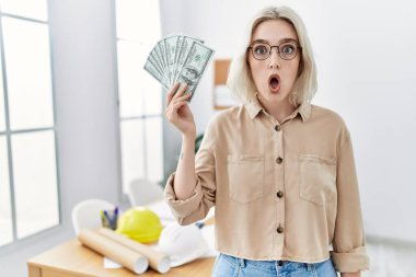Young beautiful caucasian woman at construction office holding money scared and amazed with open mouth for surprise, disbelief face 