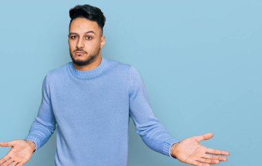 Gündelik giysiler giyen genç bir Arap, elleri ve kolları havada, kafası karışık bir ifade takınıyor. şüphe kavramı. 