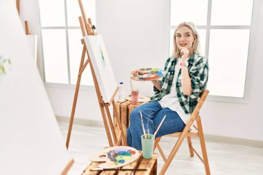 Young artist woman painting on canvas at art studio with hand on chin thinking about question, pensive expression. smiling and thoughtful face. doubt concept. 