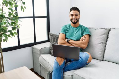 Sakallı, bilgisayar bilgisayarı kullanan genç, mutlu yüzlü, kollarını kavuşturup kameraya bakan bir adam. pozitif kişi. 