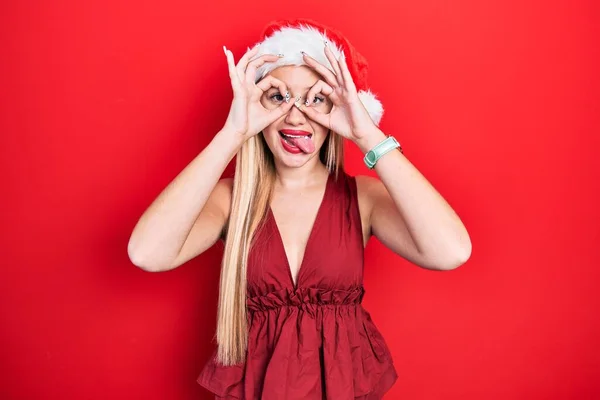 年轻的金发姑娘头戴圣诞礼帽 像双筒望远镜一样做手势 伸出舌头 用手指看着 疯狂的表达 — 图库照片