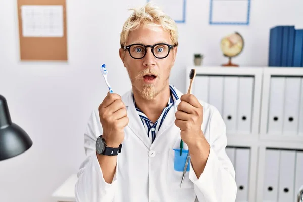 Young Blond Dentist Man Working Dentist Clinic Holding Toothbrushes Shock — Stockfoto