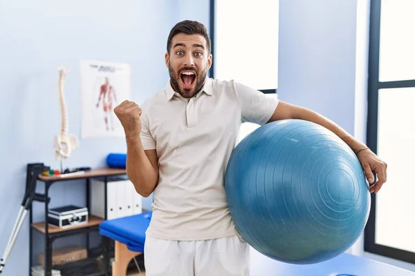 Handsome Hispanic Man Holding Pilates Ball Rehabilitation Clinic Screaming Proud — Fotografia de Stock