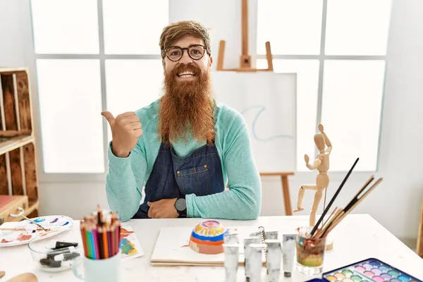 红头发的男人 留着长胡子 在艺术工作室里画着黏土碗 面带微笑 面带微笑 用大拇指指着旁边 — 图库照片