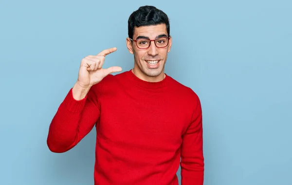 カジュアルな服や眼鏡を身に着けているハンサムなヒスパニック系の男は 指を見て カメラで小さなサイズの看板を行う手で笑顔と自信を持ってジェスチャー 概念を測定し — ストック写真