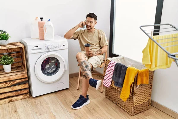 Young Hispanic Man Waiting Washing Machine Speaking Phone Laundry Room — стоковое фото