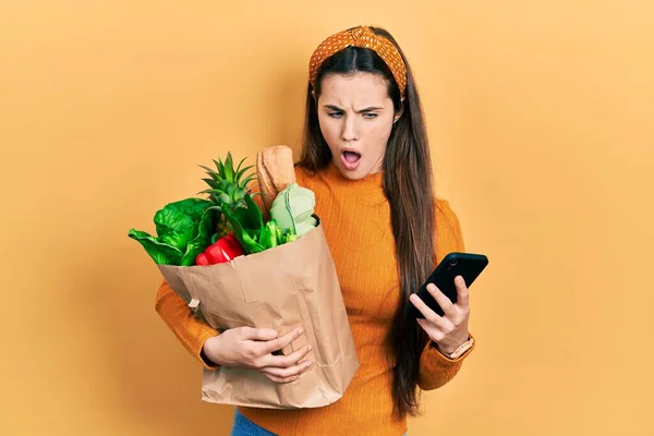 Young Brunette Teenager Holding Bag Groceries Using Smartphone Shock Face — Stock Photo, Image