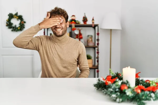 ひげを生やした若いハンサムな男がクリスマスの装飾笑顔でテーブルの上に座り 驚きのために目を覆う顔で手で笑っています 視覚障害の概念 — ストック写真