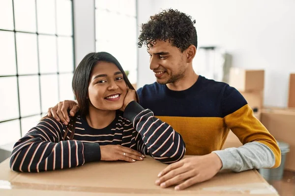 Genç Latin çift mutlu bir şekilde gülümsüyor. Yeni evlerinde karton kutuya yaslanıyorlar..