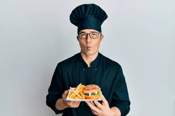 Handsome young man chef holding burger with fries making fish face with mouth and squinting eyes, crazy and comical.