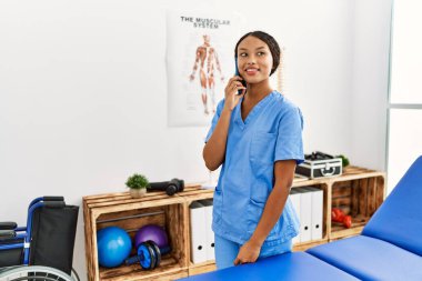 Fizyoterapist üniforması giyen genç Latin kadın klinikte akıllı telefondan konuşuyor.