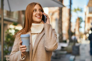 Genç kızıl saçlı kız akıllı telefonda konuşuyor. Şehirde kahve içiyor..