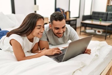 Yatakta dizüstü bilgisayar kullanarak mutlu gülümseyen genç Latin çift..