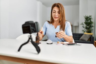 Orta yaşlı beyaz kadın makyaj dersi veriyor. Evde masanın üzerinde kamerayla oturuyor..