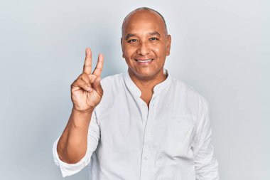 Orta yaşlı, gündelik kıyafetler giyen Latin bir adam iki numarayı gösterip işaret ederken kendine güvenen ve mutlu bir şekilde gülümsüyor.. 