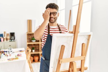 Arab young man at art studio covering one eye with hand, confident smile on face and surprise emotion. 