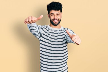 Young arab man with beard wearing casual striped sweater approving doing positive gesture with hand, thumbs up smiling and happy for success. winner gesture. 