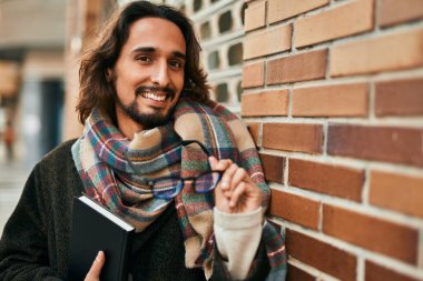 Genç İspanyol adam gülümsüyor, şehirde bardak ve kitap tutuyor..