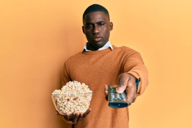 Genç Afro-Amerikan adam elinde uzaktan kumandayla patlamış mısır yiyor ve kafası karışık. şüphe kavramı. 