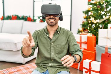 Genç İspanyol adam VR gözlük takıyor. Evdeki Noel ağacının yanında oturuyor.