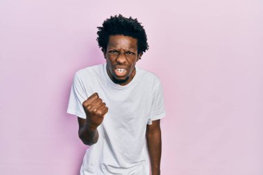 Genç Afro-Amerikalı bir adam günlük beyaz tişört giyiyor kızgın ve kızgın yumruğunu kaldırıyor ve öfkeyle bağırıyor. öfke ve agresif konsept. 