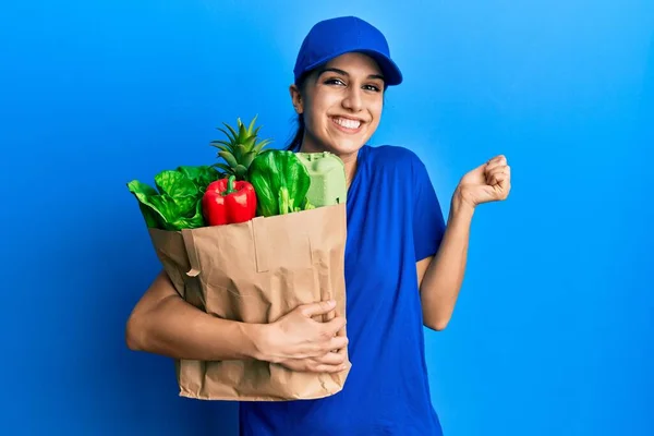 若いヒスパニック系の女性は誇りに思って叫んでスーパーマーケットから食料品と制服を着た宅配便を身に着けています 勝利と成功を祝う非常に腕を上げ興奮 — ストック写真