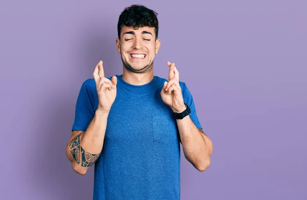 カジュアルなTシャツを着た若いヒスパニック系の男が希望と目を閉じて笑顔を交差ジェスチャー指 幸運と迷信の概念は — ストック写真