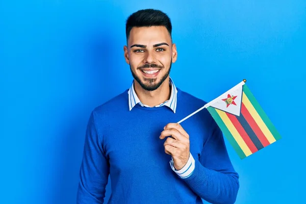 Joven Hombre Hispano Con Barba Sosteniendo Bandera Zimbabwe Luciendo Positivo —  Fotos de Stock
