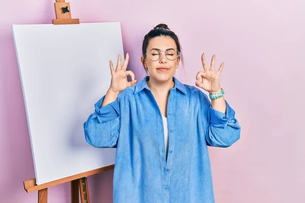 画家のイーゼルのそばに立つ若いヒスパニック系の女性は 指で瞑想ジェスチャーを行う閉じ目でリラックスして笑顔 ヨガのコンセプト — ストック写真