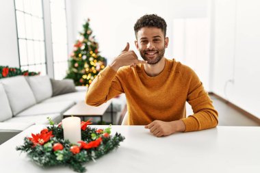 Arab young man sitting on the table by christmas tree smiling doing phone gesture with hand and fingers like talking on the telephone. communicating concepts. 