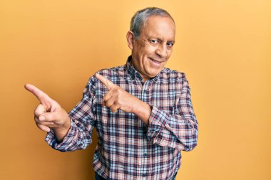 Gri saçlı yakışıklı, rahat gömlekli, gülümseyen ve kameraya bakan iki eli ve parmakları yanda olan bir adam.. 