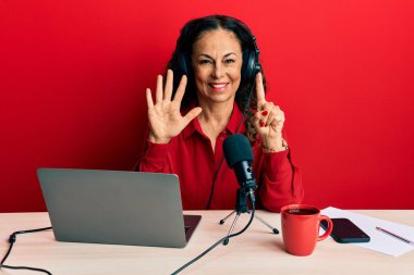 Güzel orta yaşlı bir kadın radyo stüdyosunda çalışıyor. Altıncı parmaklarını gösterip işaret ediyor. Kendinden emin ve mutlu bir şekilde gülümsüyor.. 
