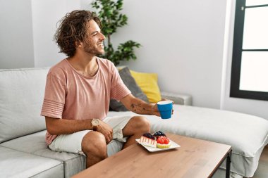 Genç İspanyol adam kahvaltısını yapıyor. Evdeki kanepede oturuyor..