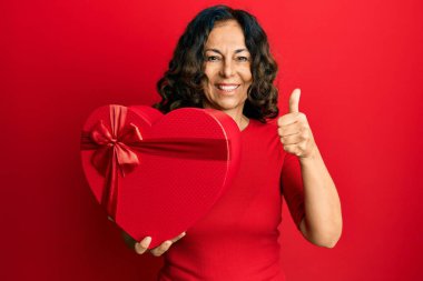 Orta yaşlı İspanyol kadın elinde sevgililer günü hediyesi, mutlu ve pozitif gülümsüyor, başparmağıyla mükemmel ve onay işareti yapıyor. 