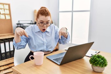 Ofiste çalışan genç kızıl saçlı kadın bilgisayarını kullanıyor. Parmaklarıyla işaret ediyor. Reklam gösteriyor, şaşırmış yüz ve ağzını açıyor. 