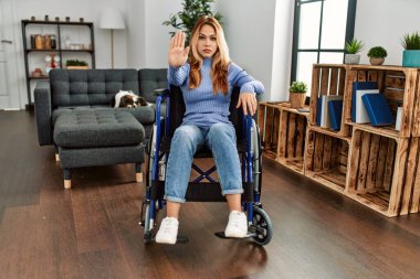 Young beautiful woman sitting on wheelchair at home doing stop sing with palm of the hand. warning expression with negative and serious gesture on the face. 