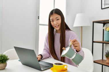 Genç Çinli kız dizüstü bilgisayar kullanıyor ve kahve içiyor. Evdeki masada oturuyor..